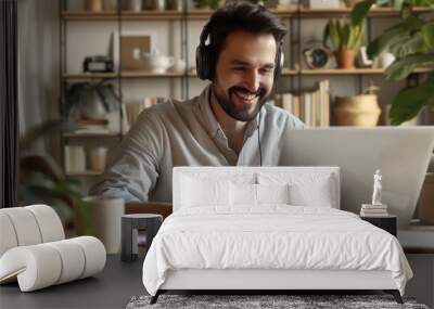 A smiling young man in headphones and glasses sits at a desk working on a laptop and making notes. Happy males in earphones watch webinars or training courses or computers and study online from home. Wall mural