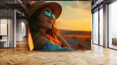 Happy woman in hat and sunglasses leaning out of the window from behind yellow car on country road, looking at beautiful landscape with sunset. She is looking in the style of summer vacation concept. Wall mural