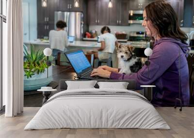 Woman working from home on laptop computer with puppy in lap while daughters study at kitchen counter in background Wall mural