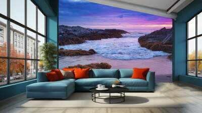 A rocky coastline with crashing waves at sunset, with a colorful sky in shades of purple, pink, and blue Wall mural