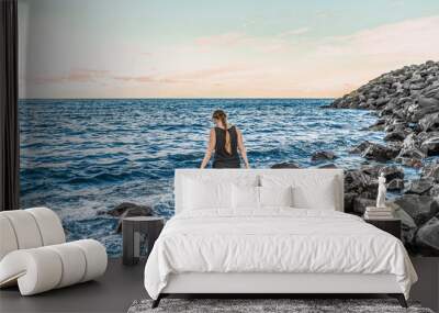 Young adult girl enters the Atlantic Ocean on the Playa Negro beach in Santa Cruz de Tenerife, Spain - view from the back. Barefoot woman in shorts and a T-shirt stands among large stones in sea water Wall mural