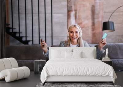 Amazed young blonde Swedish woman holding phone and credit card in headphones sitting on cozy sofa screaming happily raised hands. Online purchase discount, sale Black Friday. Successful people. Wall mural