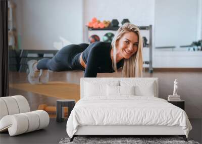 Blonde caucasian young woman in black sportswear making plank exercise on yoga mat at fitness club. Fit American girl training at gym improving endurance. Sport, healthy lifestyle. Sport  and health. Wall mural