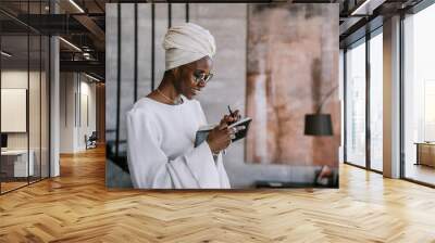 Cheerful African woman in white turban, white dress, glasses holding planner writing notes cute smiling satisfied by her life, standing at home. Thankful  afro american female in traditional clothes. Wall mural