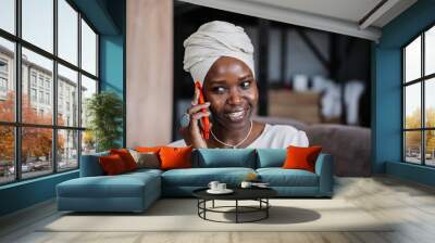 Cheerful African young woman in turban sitting on sofa talking by phone toothy smiles looks aside. Happy businesswoman in traditional african clothes discussing with partner using sell phone. Wall mural