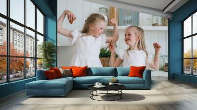 Cheerful blonde little sisters in white dresses looking at each other smiling showing biceps muscle at kitchen, having fun. Cute girl shows cookie to sister. Childhood, domestic leisure. Wall mural