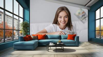 Cheerful caucasian girl sitting on bed with money throwing dollar banknotes at camera toothy smiling. Gorgeous swedish blonde young confident woman got business profit. Healthy life, financial career. Wall mural