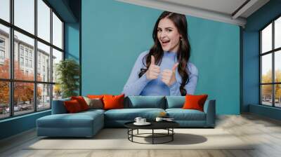 Cheerful young brunette hispanic woman in lilac sweater shows thumb up gesture by both hands looks at camera excited by discount, sale, promo over turquoise studio backdrop with empty space. Wall mural