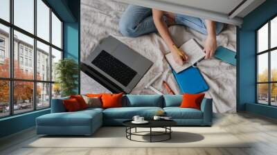 Cropped shoot of girl writing in diary sitting on bed with laptop phone pen, remote learning home. Top view on remote working young woman planning agenda at bedroom. Education, business people. Gadget Wall mural