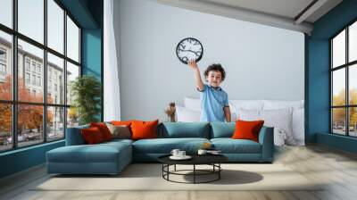 Cute caucasian boy in blue polo holding wall clock standing on bed, early morning getting up. Time management concept. Curly Spanish kid looks at camera. Kid asks to spend time with parents. Baby boy Wall mural