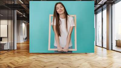 Cute cheerful young woman in white t-shirt and jeans holds frame for Canvas smiling wide stands over turquoise studio backdrop with empty space. Attractive Italian girl got into wooden frame. Mockup Wall mural