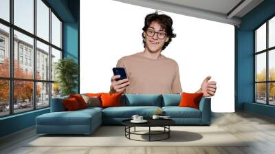 Excited teen in glasses toothy smiles holds phone, amazed by message. Young brunet guy in beige t-shirt, glasses received great news, entered university. American boy isolated, transparent background Wall mural