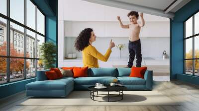 Little Spanish curly boy in blue pants standing on a table showing his biceps, smiling, her mom looking at him with proud gesturing thumb up at kitchen. Healthy kid rising fists up in a winner gesture Wall mural