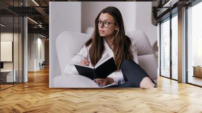 Purposeful smart businesswoman in glasses sitting at cozy chair at home holds diary looks aside, planning agenda. Attractive hispanic brunette entrepreneur remote working. Student writing in notebook. Wall mural