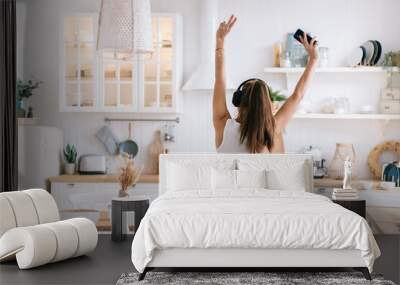 rear view of a positive girl in jeans dancing in the kitchen at home using headphones and a phone that raised her hands up shows a victory sign a fun weekend at home Wall mural