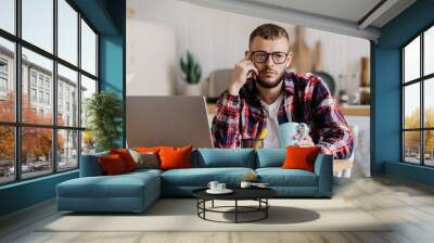 Sad Scandinavian young man in plaid shirt and glasses sits at desk with laptop holds cup of coffee with frustrated face stares away feels loneliness. Overworked caucasian guy tired. Failure, mistake. Wall mural