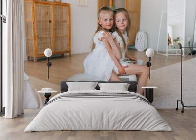 Two little blonde Swedish sisters sitting on toy car at living room. Caucasian girls playing together wearing white dresses. Childhood, family leisure. Satisfied girl happy to spend time with sister. Wall mural