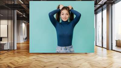 Young adult pretty woman in blue sweater, jeans looks at camera, holding her hair, making two ponytails over turquoise studio backdrop with empty space. Mockup. Cute Swedish girl with healthy hair. Wall mural