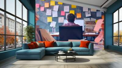 Man sitting at a desk with a stack of books, notes on the wall, working on a project or studying Wall mural