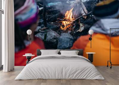 : Two people in warm clothing sit by a campfire in the wilderness, surrounded by rocks and sticks, enjoying the warmth and companionship. Wall mural