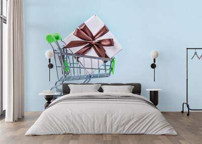 Gift box with brown bow in miniature shopping cart on pastel blue background with copy space. Wall mural