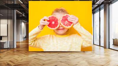 Smiling child girl holding two halfs of yellow grapefruit citrus fruit in hands, covering her eyes, on yellow. Healthy diet nutrition. Happiness fun concept Wall mural