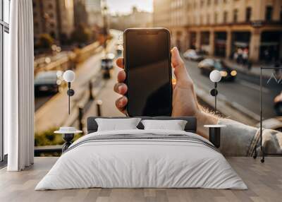 Male hand holding a blank smartphone mockup in front of a blurred city background . Copy space Wall mural