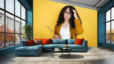 African american business woman with paperwork in hands over yellow background confuse and wonder about question. Uncertain with doubt, thinking with hand on head Wall mural