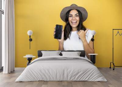 Beautiful woman wearing casual white t-shirt and a hat standing over yellow background smiling and pointing to the copy space Wall mural