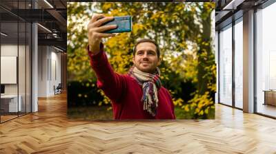 Photo of a young and attractive man in the park during autumn season taking a selfie with his phone. He is wearing a scarf and is smiling. Surrounded by trees with yellow, green and orange colours. Wall mural