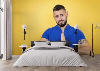 Handsome man with beard wearing blue polo shirt over yellow background begging and praying with hands together with hope expression on face very emotional and worried Wall mural