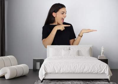 Young beautiful woman standing over white isolated background surprised, smiling and showing something that is on her hand Wall mural