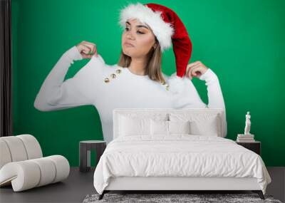 Young beautiful woman wearing a Santa hat and white sweater over green background holding her t-shirt with a successful expression Wall mural