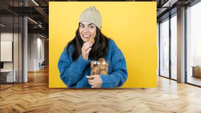 Young beautiful woman wearing blue casual sweater and wool hat holding chocolate chips cookies jar feeling guilty and taking a cookie Wall mural