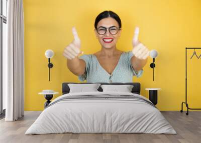 Young beautiful woman wearing glasses standing over isolated yellow doing happy thumbs up gesture with hands. Approving expression looking at the camera showing happiness background Wall mural