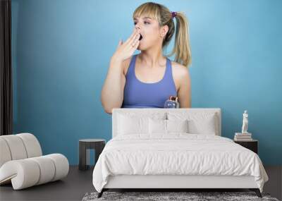 Young beautiful woman wearing sportswear holding a bottle of water over isolated blue background bored yawning tired covering mouth with hand. Restless and sleepiness. Wall mural