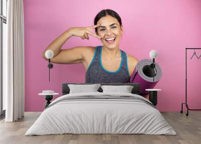 Young beautiful woman wearing sportswear over isolated pink background Doing peace symbol with fingers over face, smiling cheerful showing victory. Holding a splinter in her hand. Wall mural