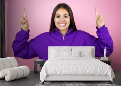 Young beautiful woman wearing sweatshirt over isolated pink background smiling, looking at the camera and pointing up with fingers and raised arms Wall mural