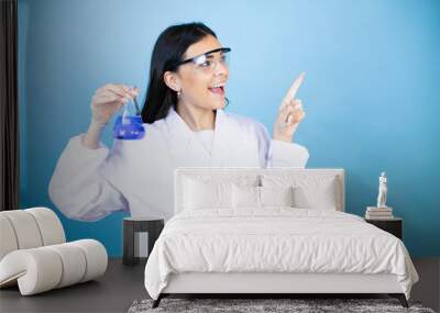 Young brunette woman wearing scientist uniform holding test tube over isolated blue background amazed and pointing with hand and finger to the side Wall mural