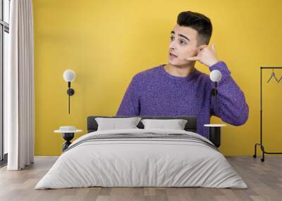 Young caucasian man over isolated yellow background confused doing phone gesture with hand and fingers like talking on the telephone Wall mural