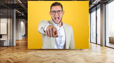 Young handsome businessman wearing suit over isolated yellow background laughing at you, pointing finger to the camera with hand over body, shame expression Wall mural