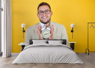 Young handsome businessman wearing suit over isolated yellow background smiling and holding a clock Wall mural