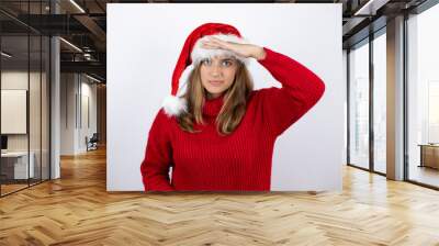 Young pretty blonde woman wearing a red casual sweater and a christmas hat over white background very happy and smiling looking far away with hand over head. searching concept. Wall mural