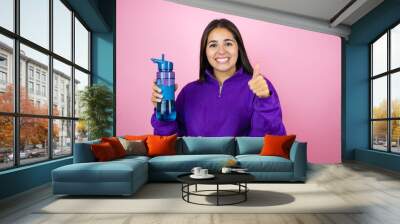 Young woman wearing sweatshirt holding a water bottle over isolated pink background success sign doing positive gesture with hand, thumb up smiling and happy. cheerful expression and winner gesture Wall mural