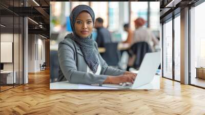 A hijab woman wearing a black and white scarf sits at a desk with a laptop Wall mural