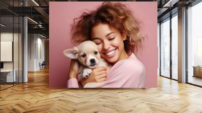 Happy young woman with curly hair hugging cute puppy, smiling as pet owner holding dog with love and care in studio, isolated on pink background Wall mural