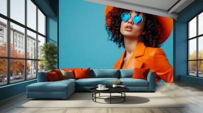 Portrait of a glamorous female model in sunglasses, a hat and a bright orange leather suit, with curly hair on a blue background in a gradient style, with copy space for text. Style and high fashion Wall mural