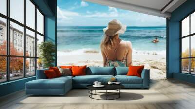 A woman in a summer dress and sun hat sitting on the sandy beach, gazing at the tranquil ocean while a swimmer enjoys the water in the background Wall mural