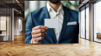 Man in a suit holding a white blank business card, standing in a professional setting, offering his contact information. Wall mural