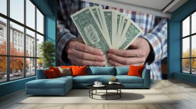 Close-up on the hands of a young man counting money into a bundle of one dollar bills arranged in a fan against the background of a plaid shirt Wall mural